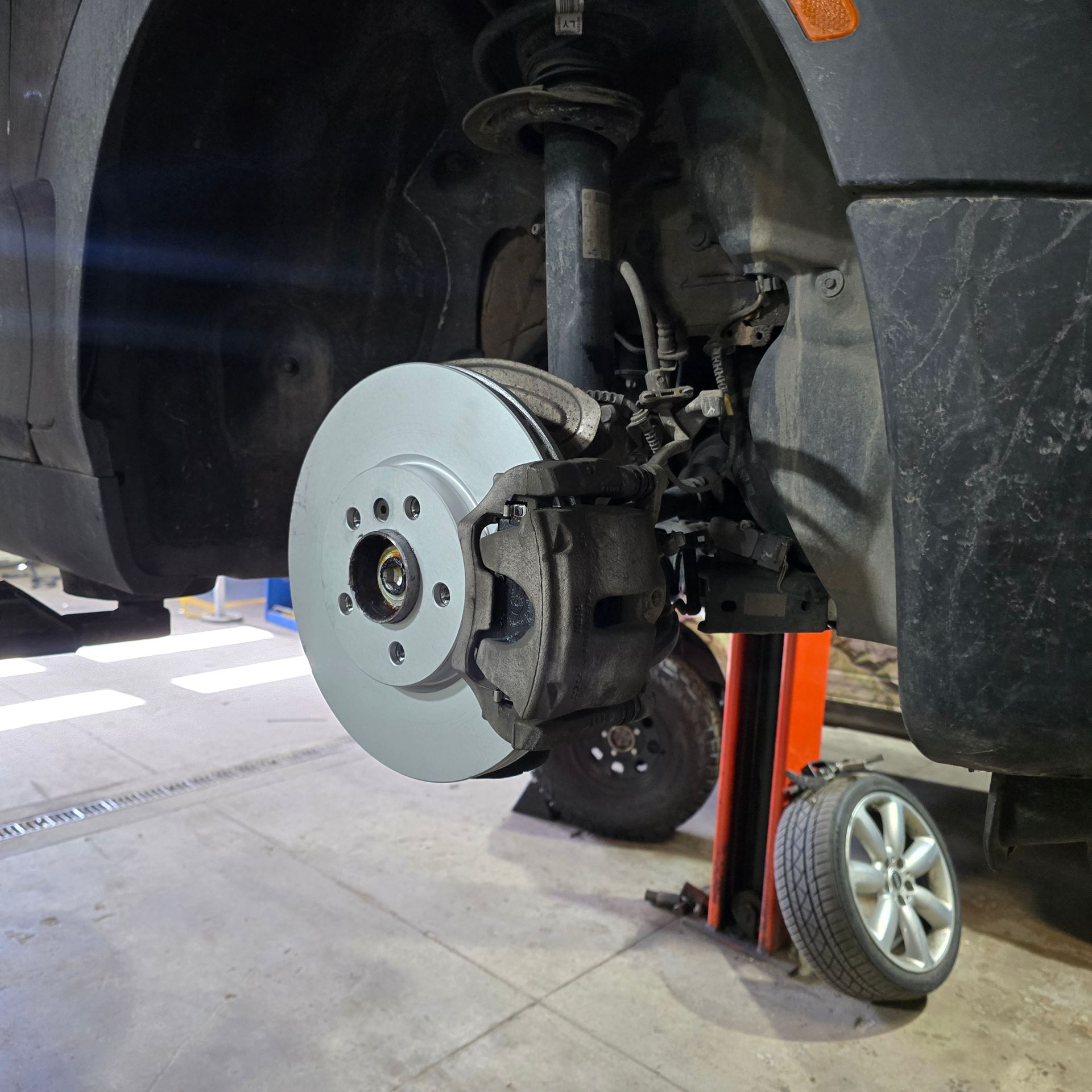 Car's front brake disc and caliper are visible during repair. Wheel is removed. Workshop setting.