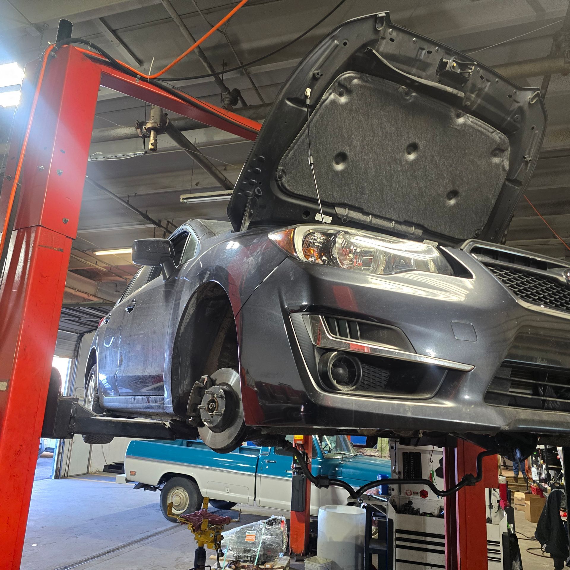Gray car on a lift in a garage with hood open, brake removed. A blue truck is visible in the background.