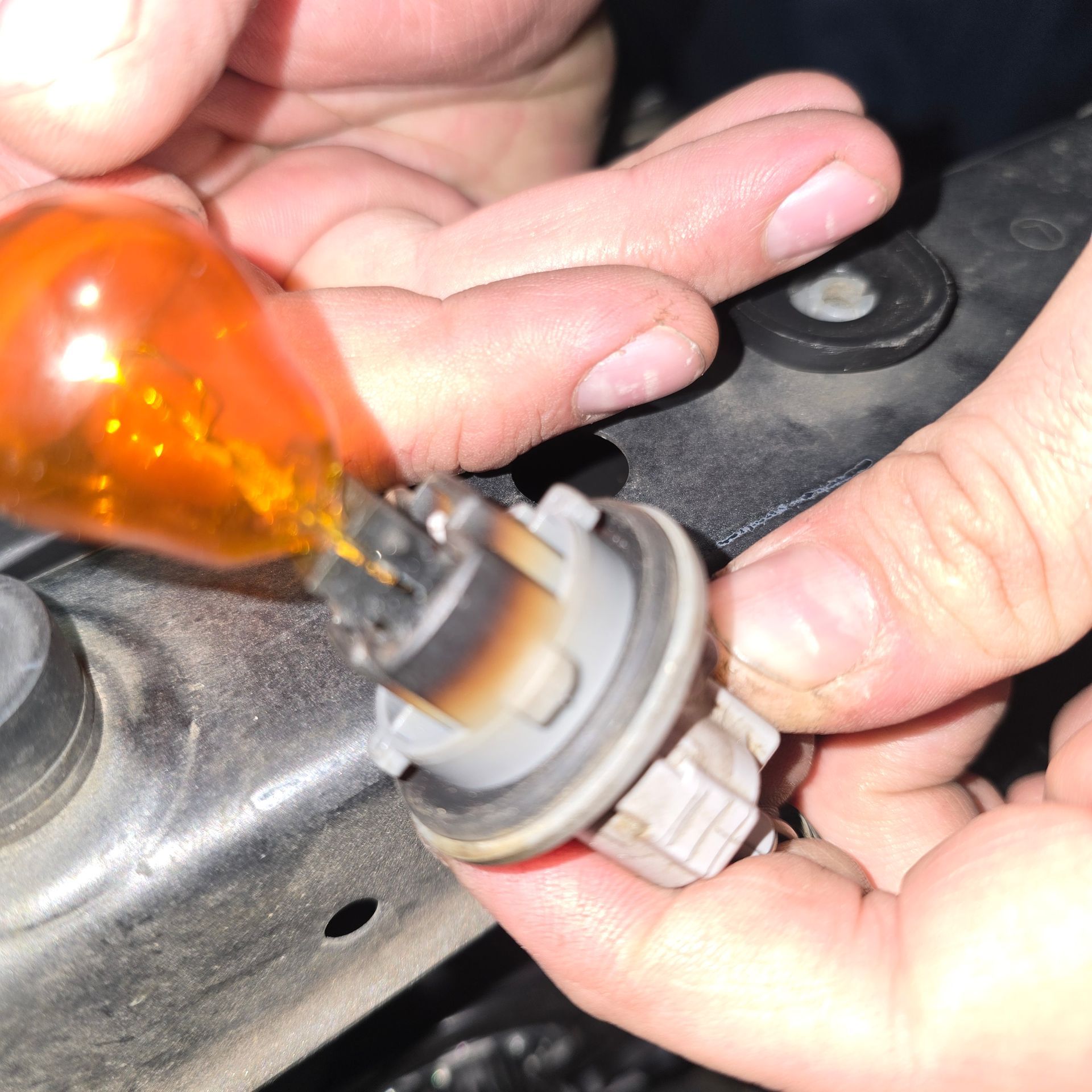 Hands holding an orange light bulb and its socket near a car headlight assembly.