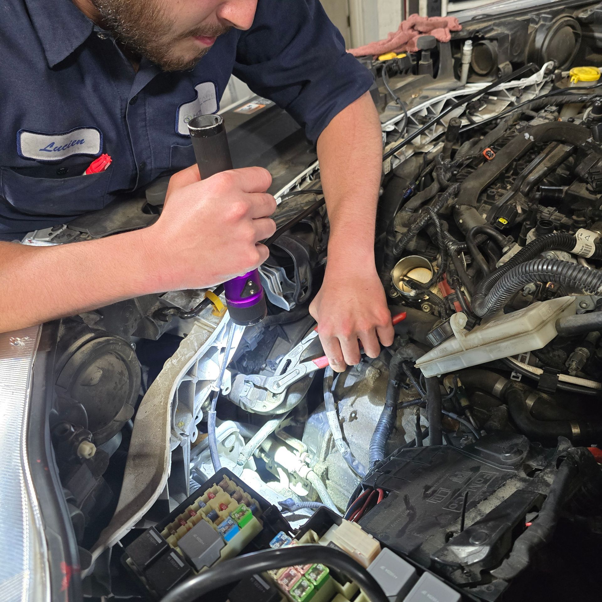 Mechanic inspecting car engine with flashlight. He's wearing a blue work shirt, looking intently. Engine bay visible.