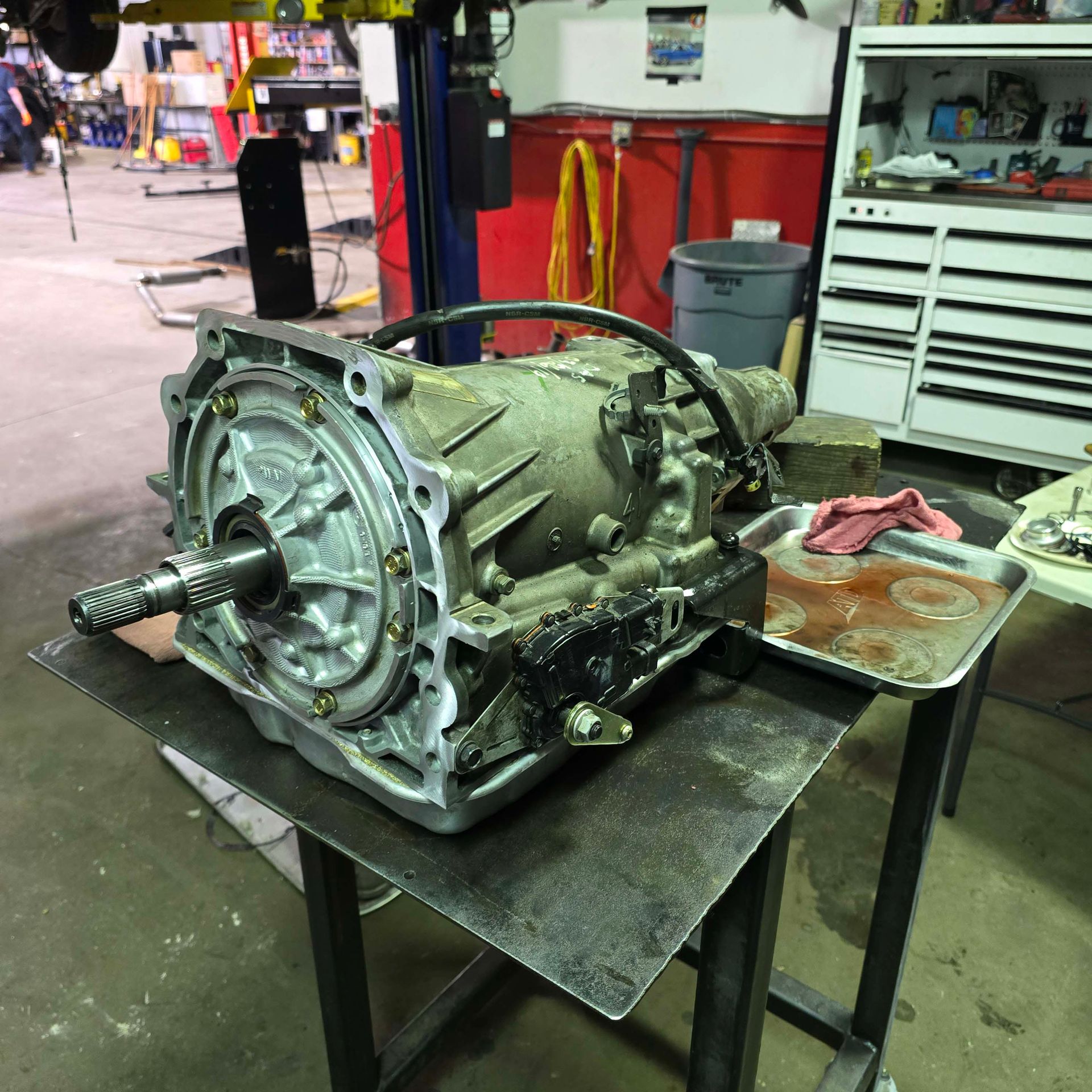 An automotive transmission rests on a metal workbench in a repair shop with a drip pan collecting fluid underneath.