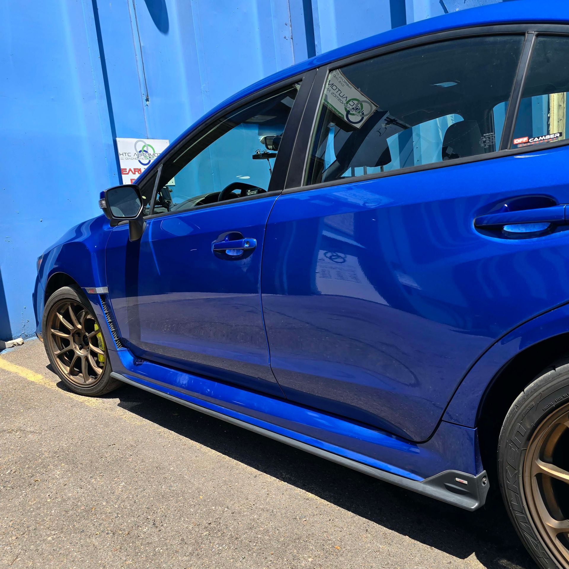 A side view of a bright blue sport sedan with dark bronze wheels, parked against a blue wall.