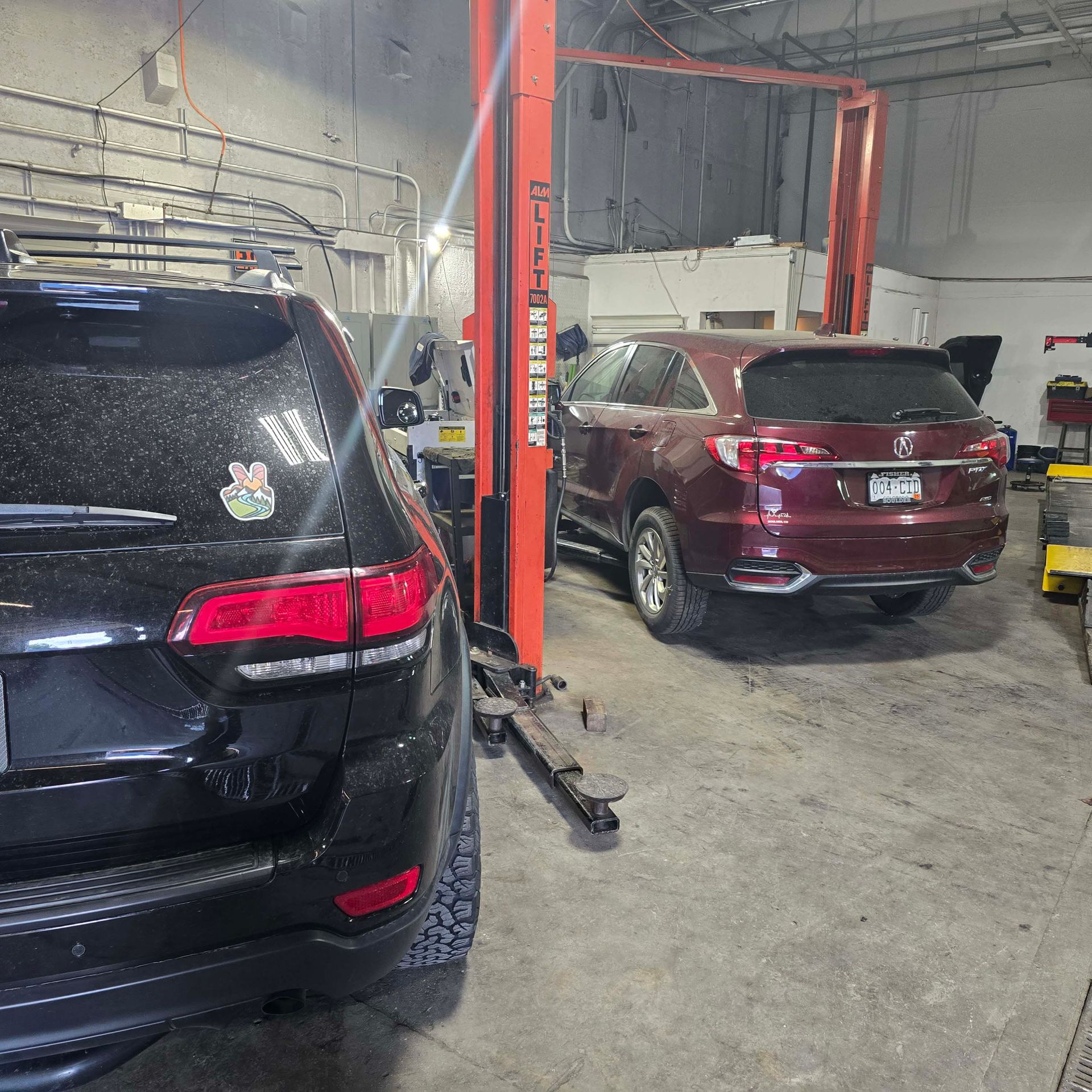 A black SUV in the foreground and a maroon SUV on a car lift inside an automotive repair shop.