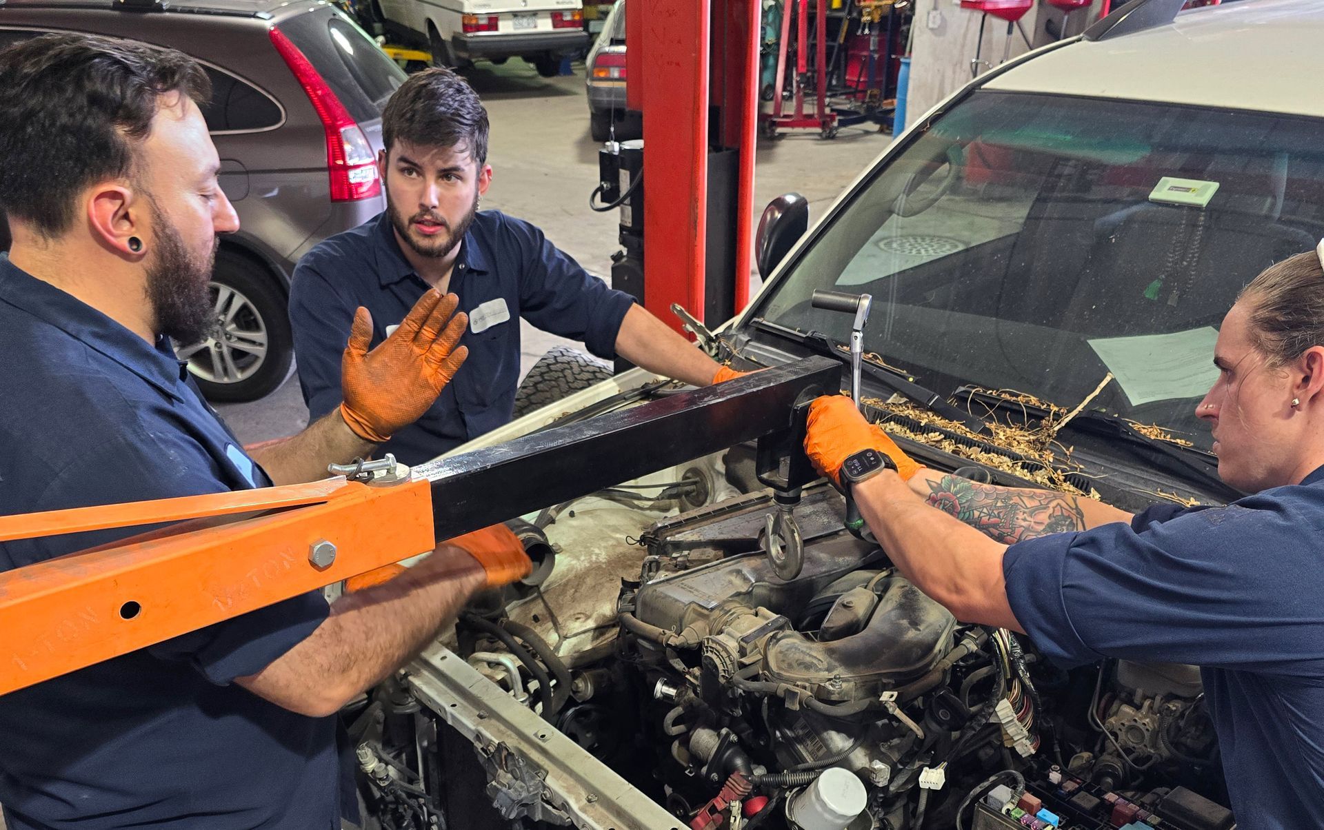 Three mechanics working on a car engine in a garage. One operates a hoist while the others assist, wearing gloves.