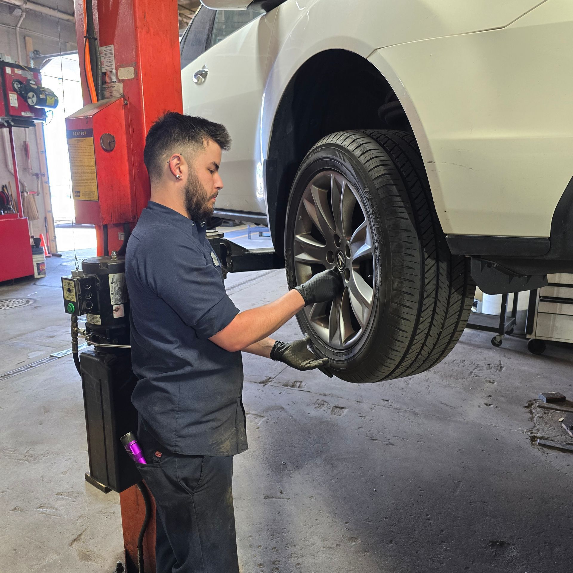 Mechanic removes a car tire in a garage, using a lift. The car is white.