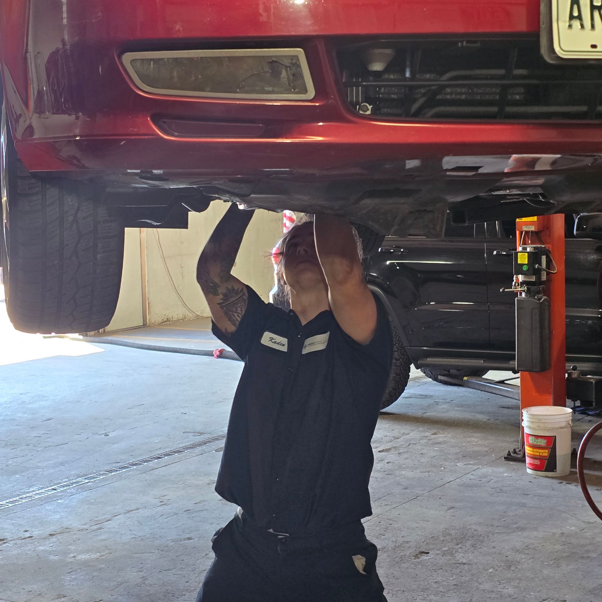 Mechanic under a red car, working on the undercarriage. They're wearing a black uniform and have tattoos on their arms.