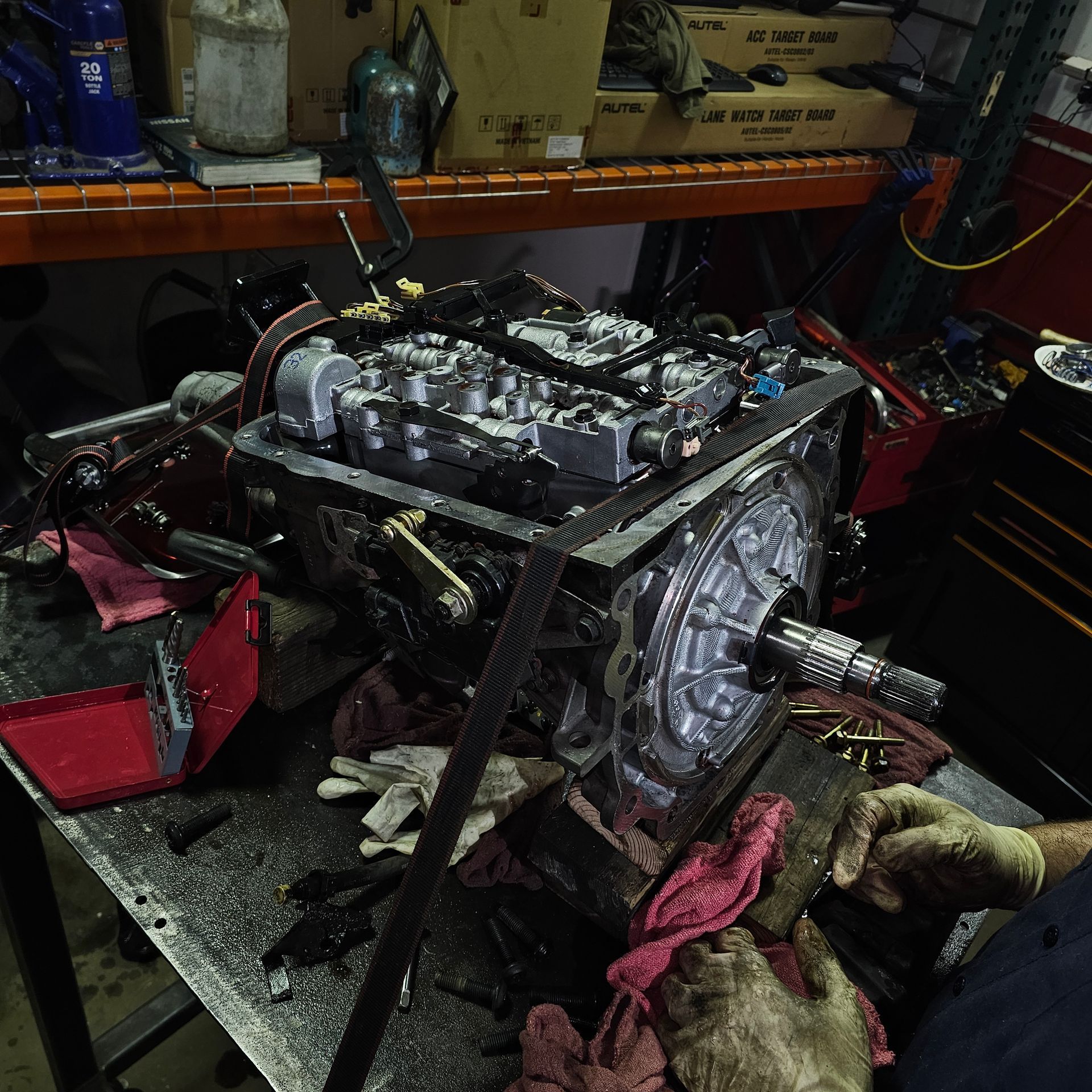 Mechanic examines a transmission on a lift in a garage, wearing a cap and blue overalls.