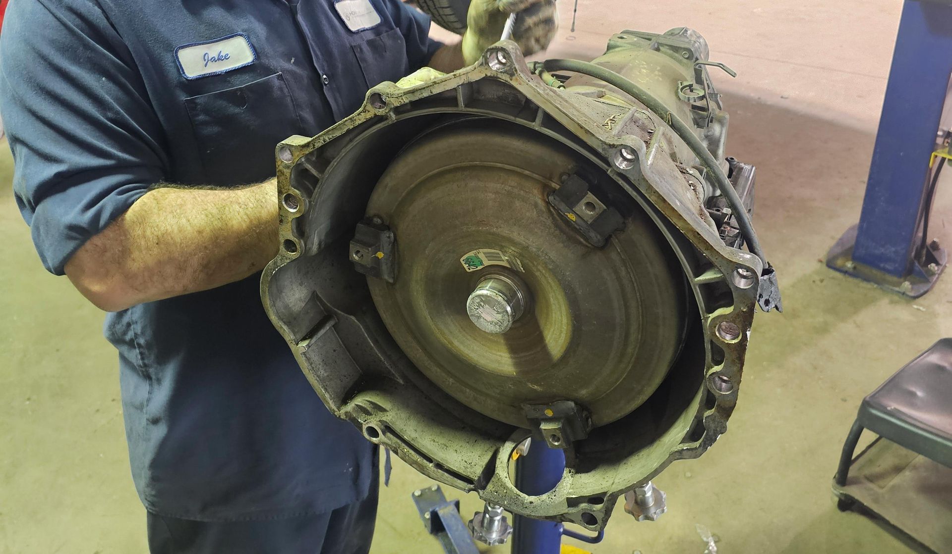 Mechanic holding an automatic transmission with a torque converter visible. Workshop setting.