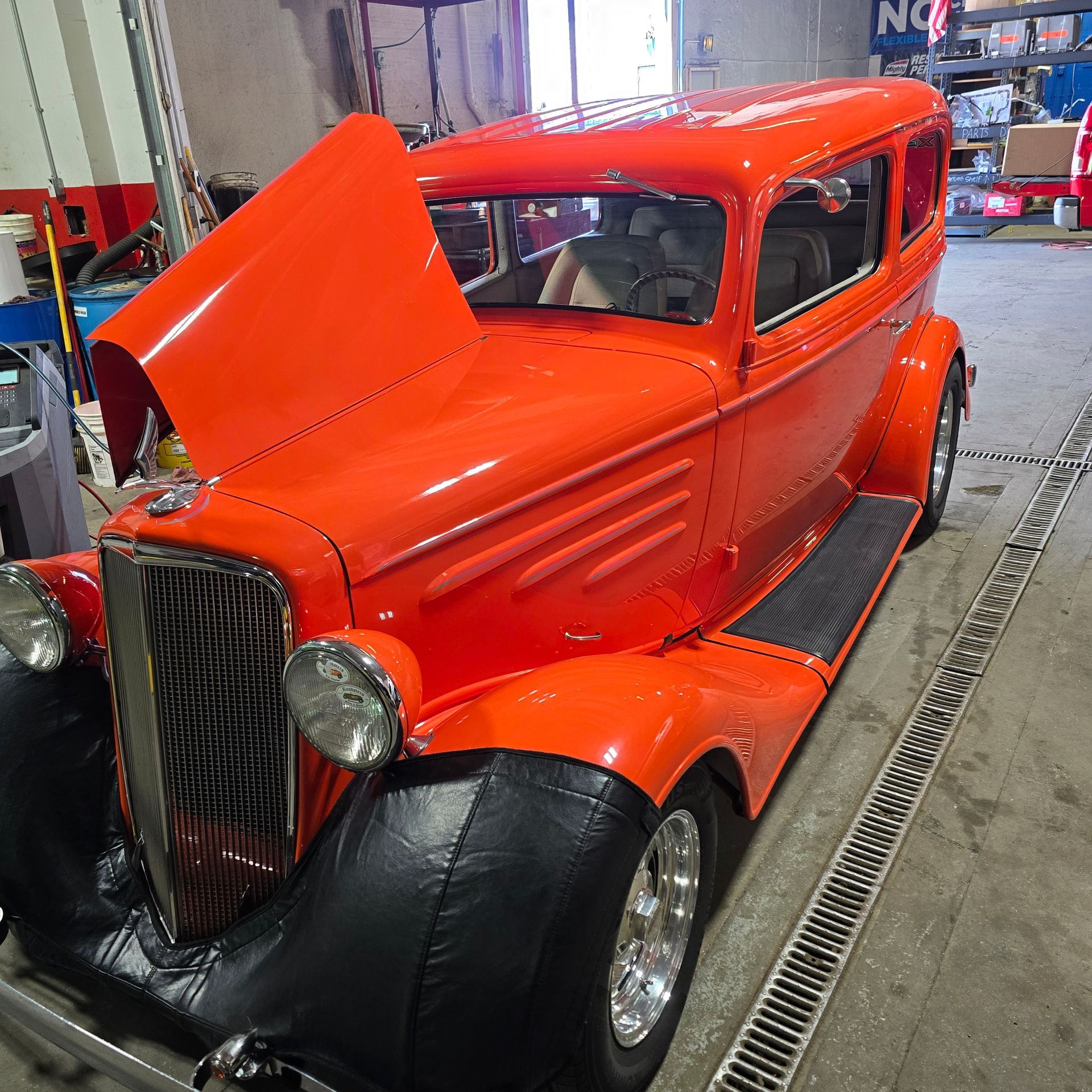 Bright orange classic car with hood open in a garage, black fender covers, chrome wheels.