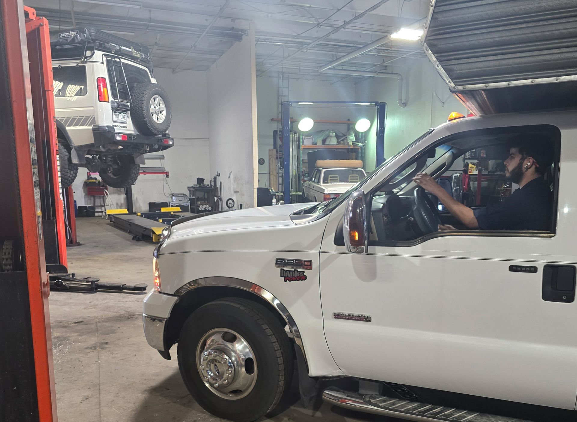 A mechanic in a white truck drives into a shop with a vehicle on a lift in the background.
