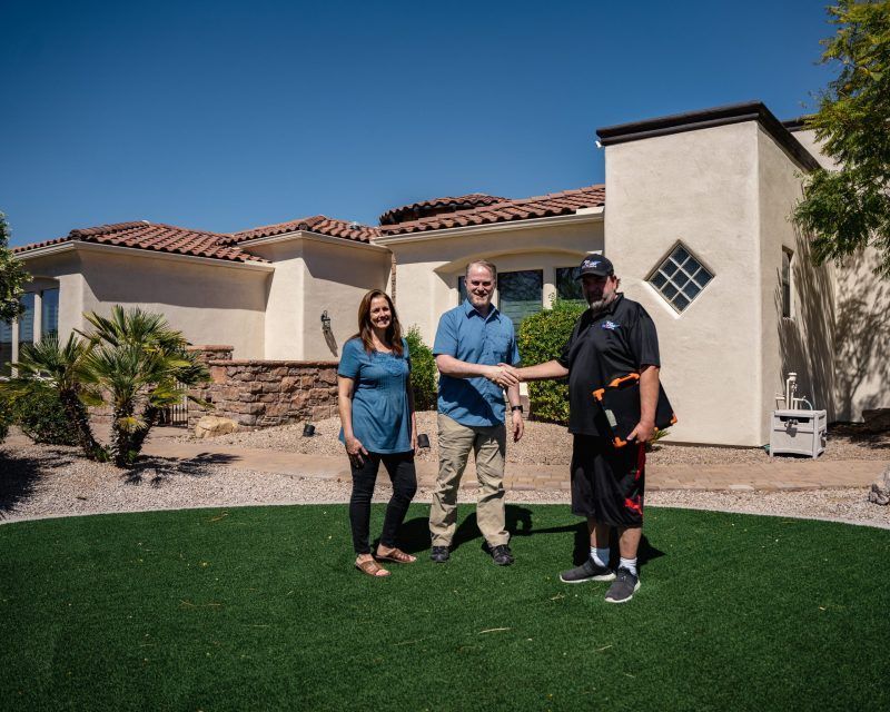 Three people standing on a lawn in front of a modern house, smiling and holding a key together.