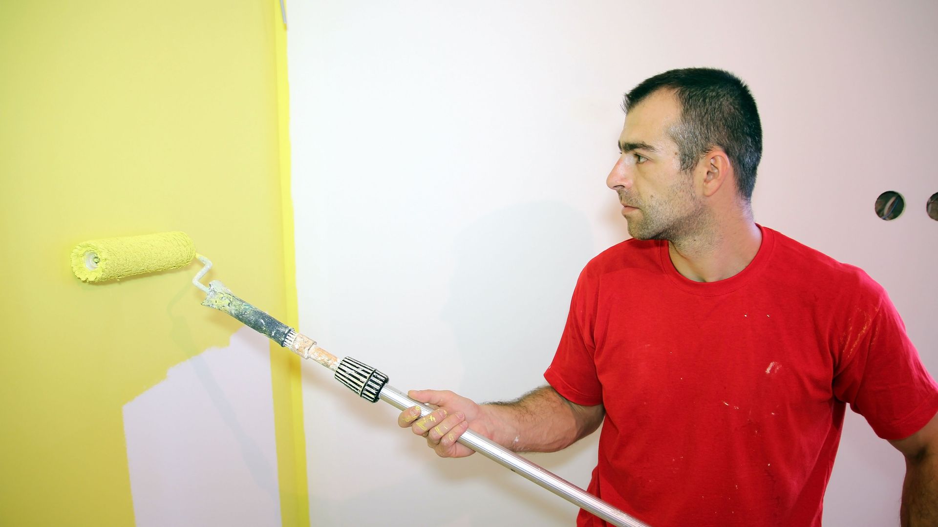 Man in red shirt painting a yellow wall with a roller, standing beside a white wall