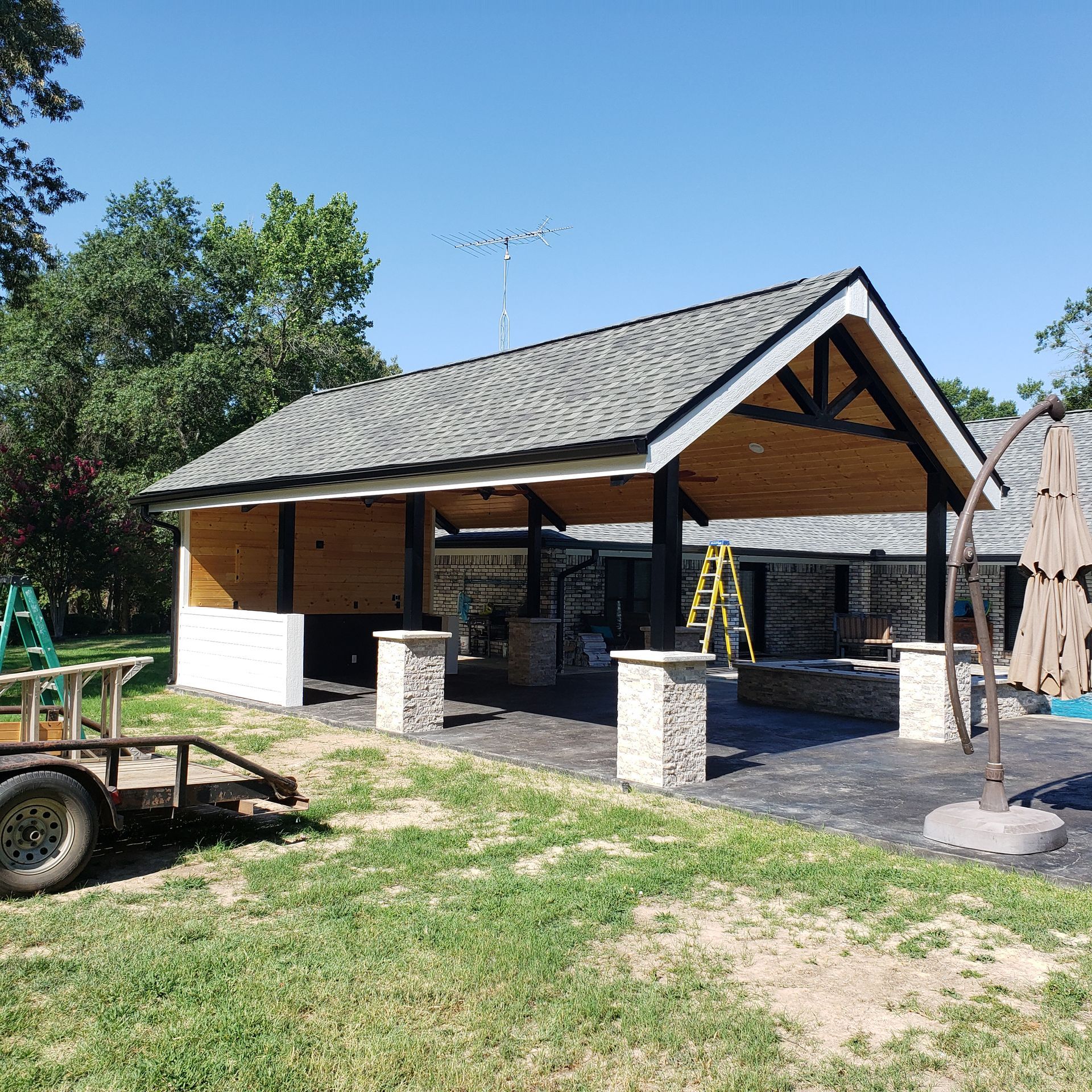 Patio with stone pillars, grey roof, wooden ceiling. Trailer and ladder on grass. Blue sky.