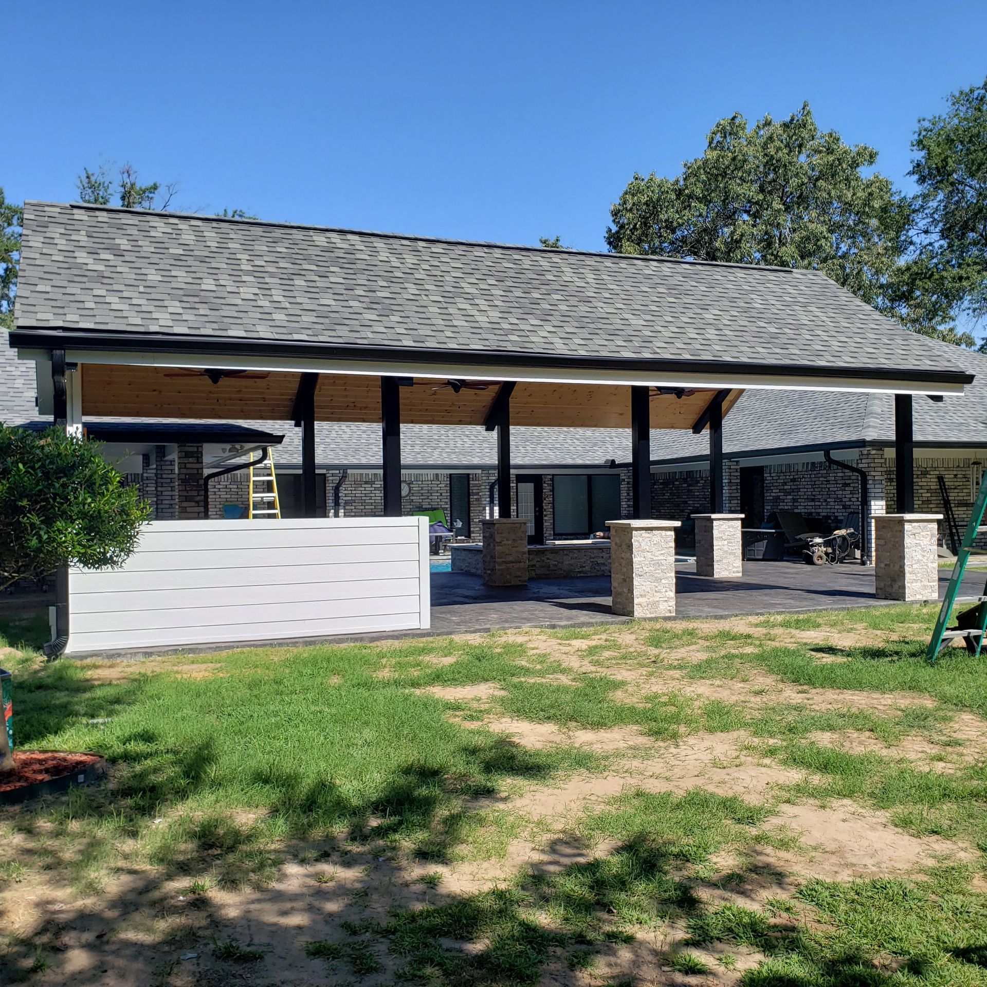 Outdoor patio with gray roof, black columns, beige pillars, and a white fence, on a sunny day.