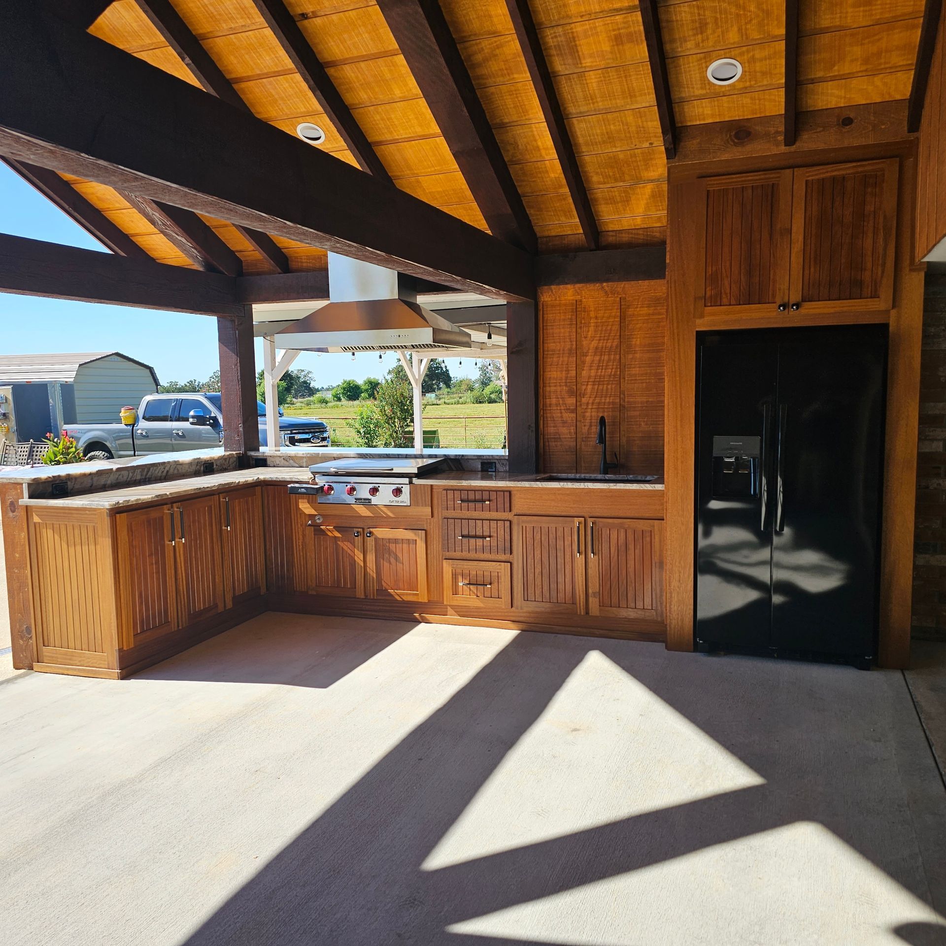 Outdoor kitchen with wooden cabinets, stainless steel appliances, and concrete patio under a canopy.