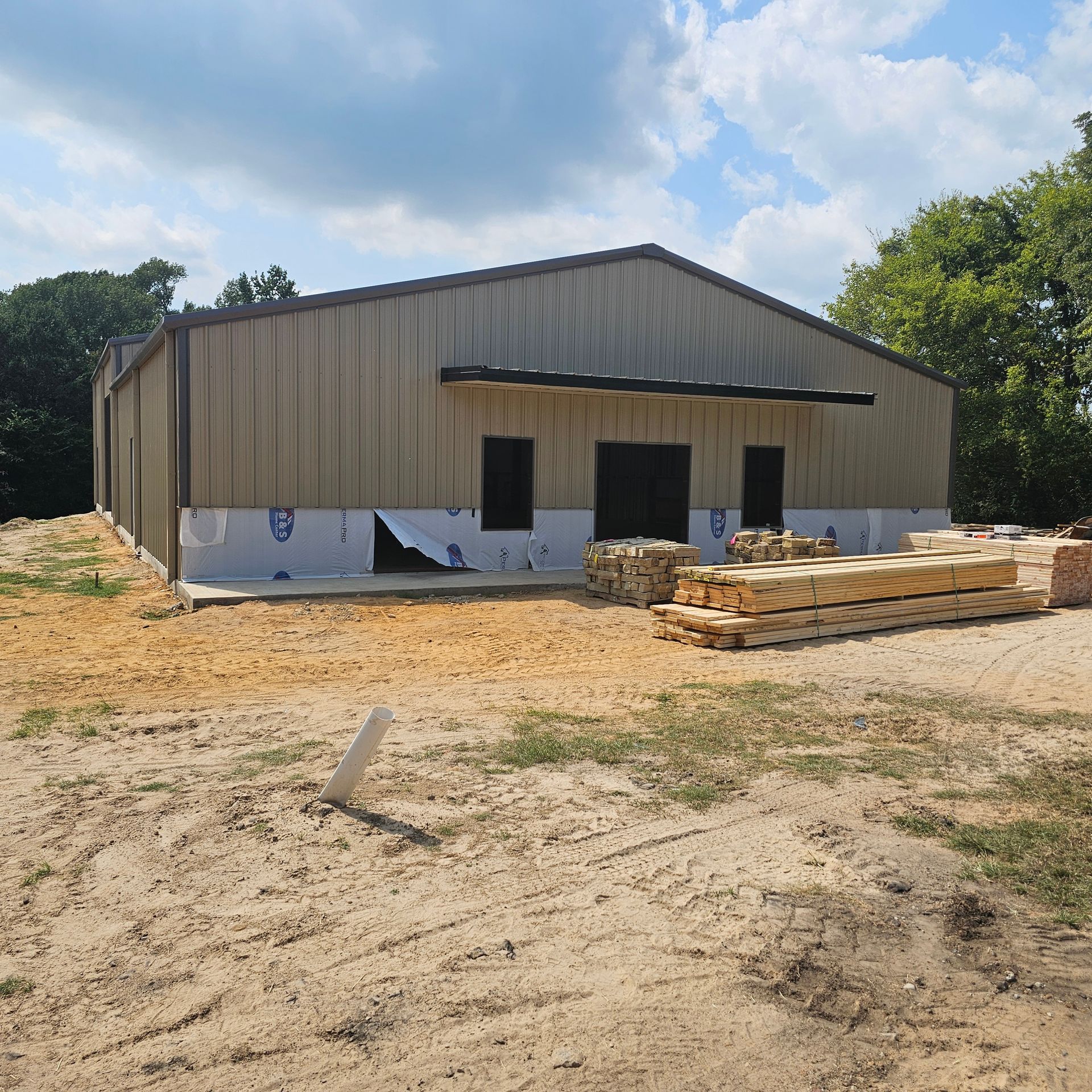 Metal building under construction on a dirt lot, with wood beams stacked in the foreground.