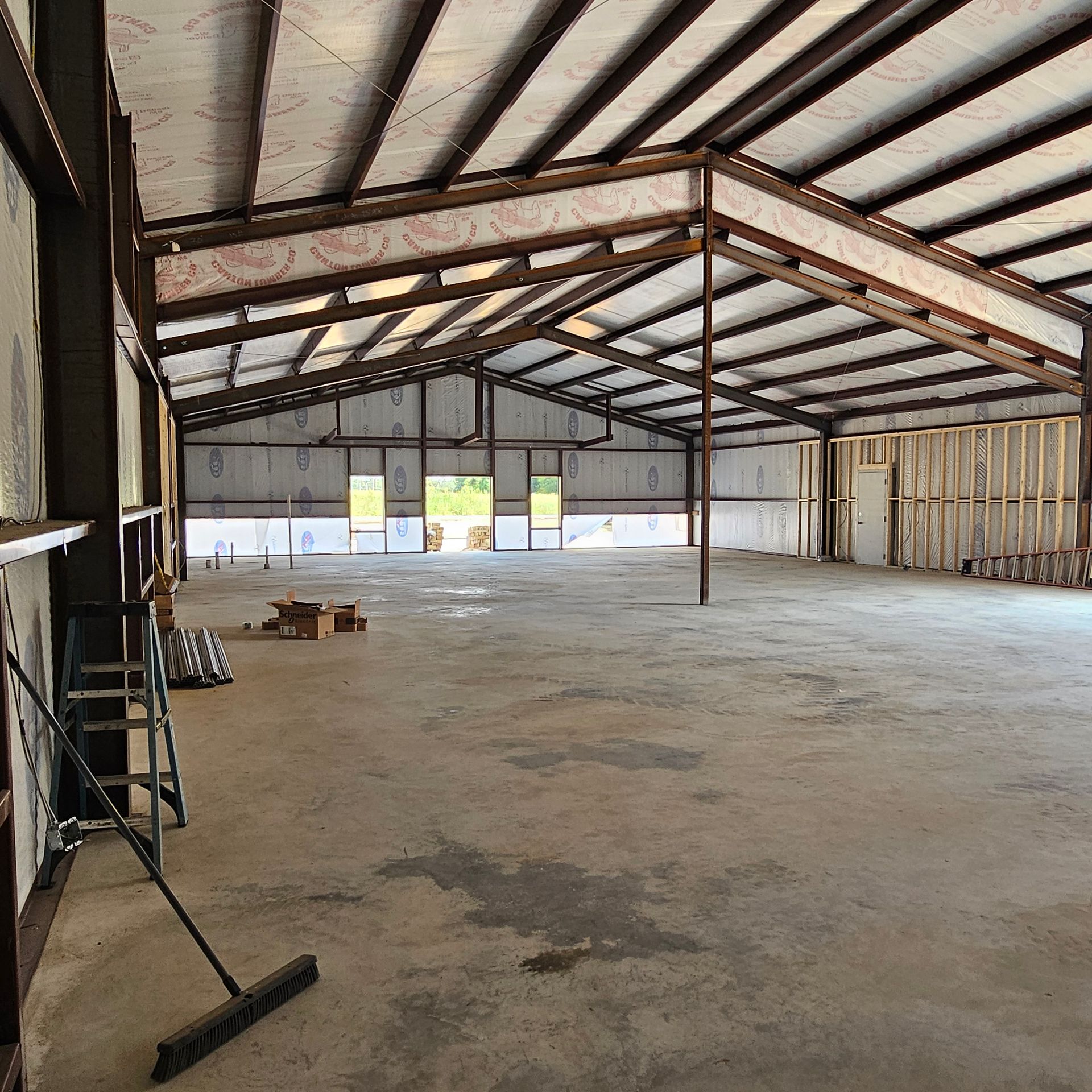 Interior of a large unfinished metal building. Concrete floor, steel beams, and a partially framed wall.