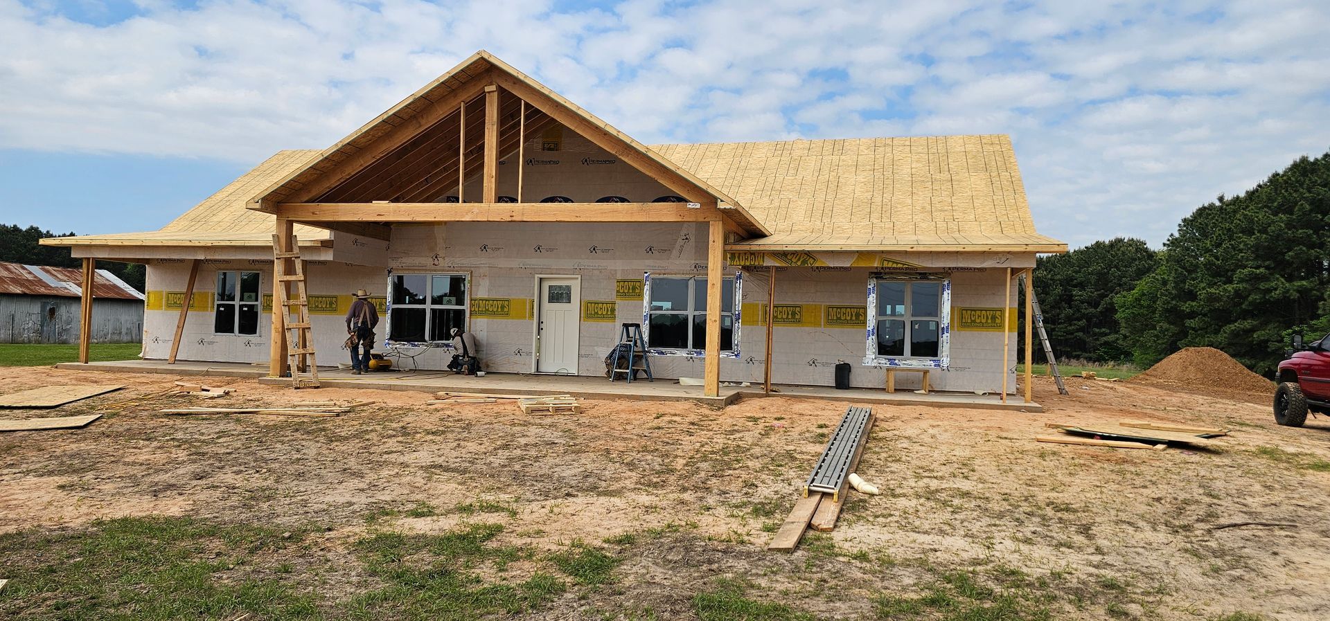 House under construction with exposed framing, plywood roof, and vapor barrier. Dirt lot with piles of materials.