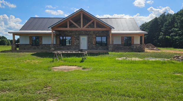 New house with stone and wood accents, metal roof, and large porch. Located in a grassy field on a sunny day.
