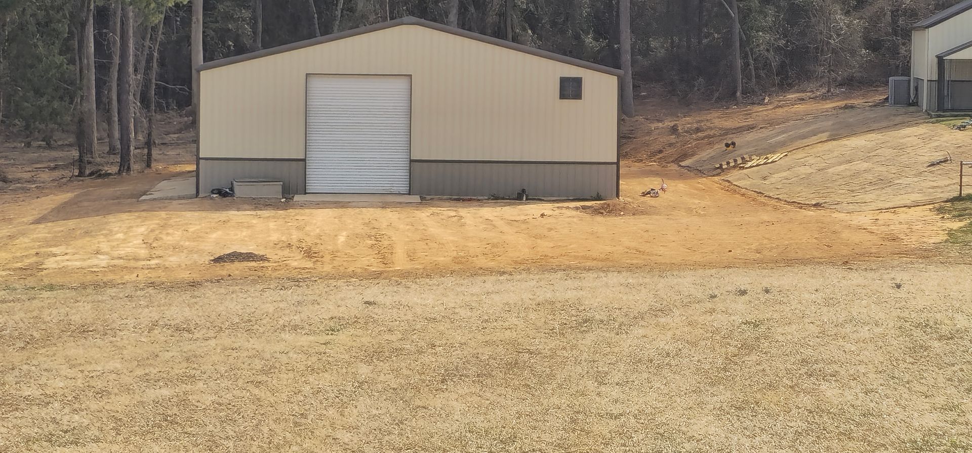 Tan building with a garage door, set in a yard with gravel and dry grass. Trees are in the background.