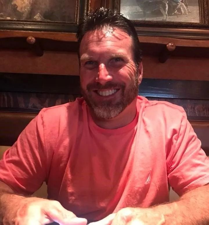 Man with a beard smiles, wearing a pink shirt, seated at a table.