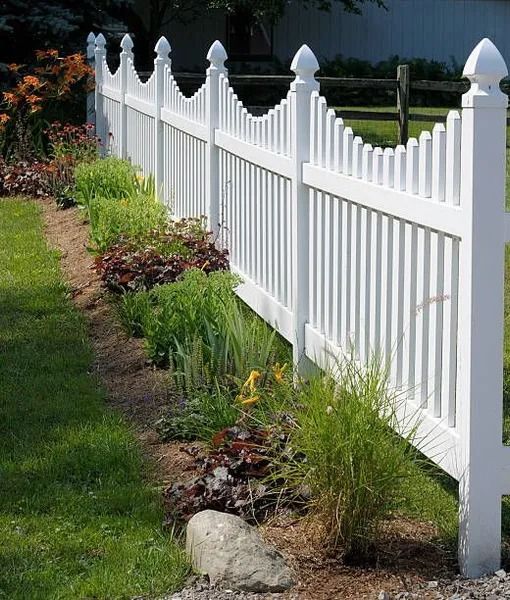 White picket fence with decorative finials along a flower bed bordering a grassy lawn.