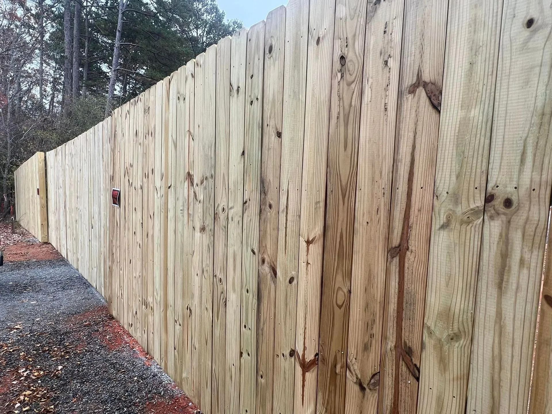 Wooden fence, vertical planks, in an outdoor setting with trees and gravel.