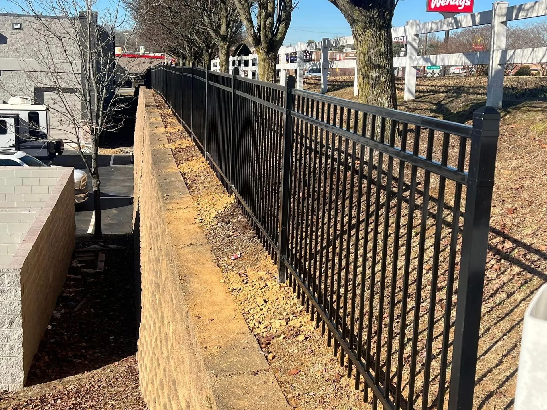 Black metal fence atop a retaining wall, with a street and businesses visible in the background.