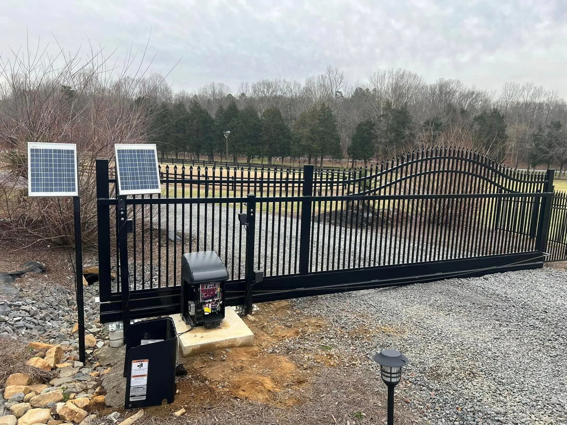 Black metal automatic gate with solar panels in a rural setting.