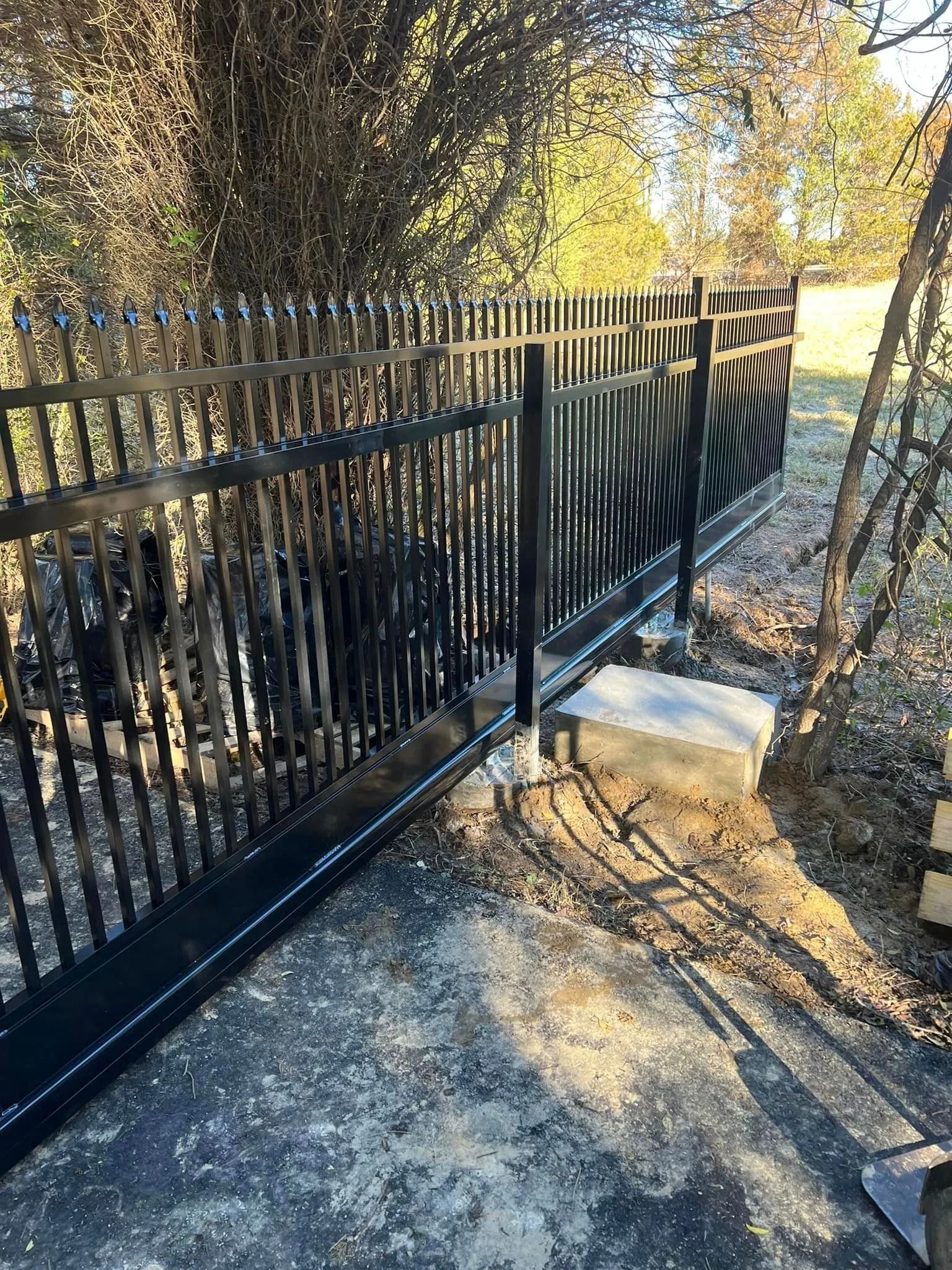 Black metal fence with gate, concrete base, setting near trees.