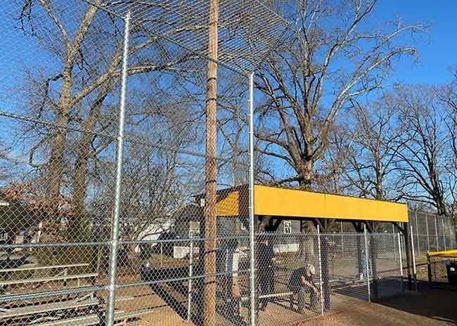 Baseball field with chain-link fence, bleachers, and dugout. Person seated on bench. Trees and blue sky in background.