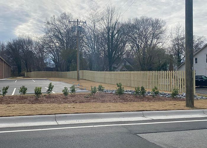 Wooden fence lines a landscaped area with shrubs and a parking lot against a background of trees and a cloudy sky.