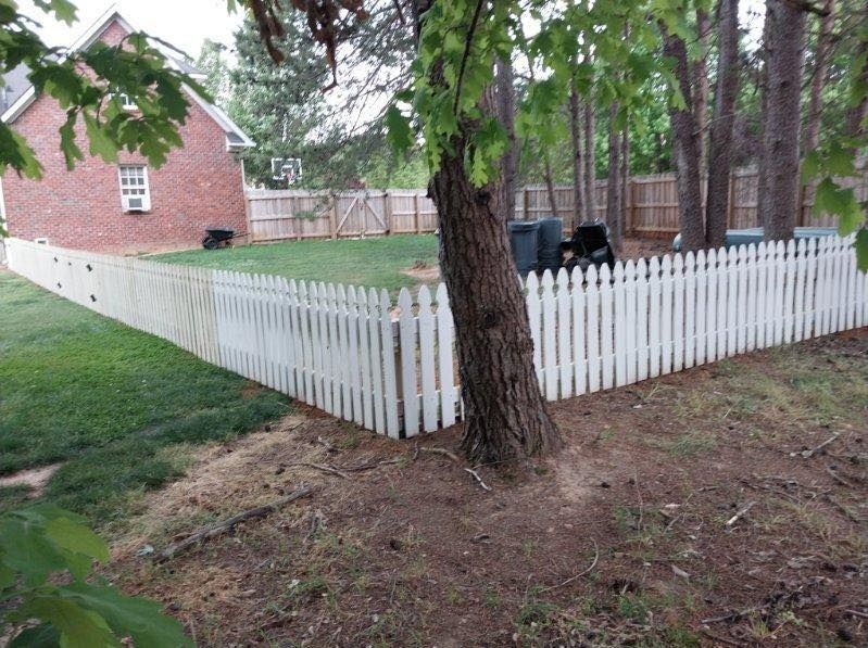 White picket fence surrounds a grassy backyard, with a brick house and trees.