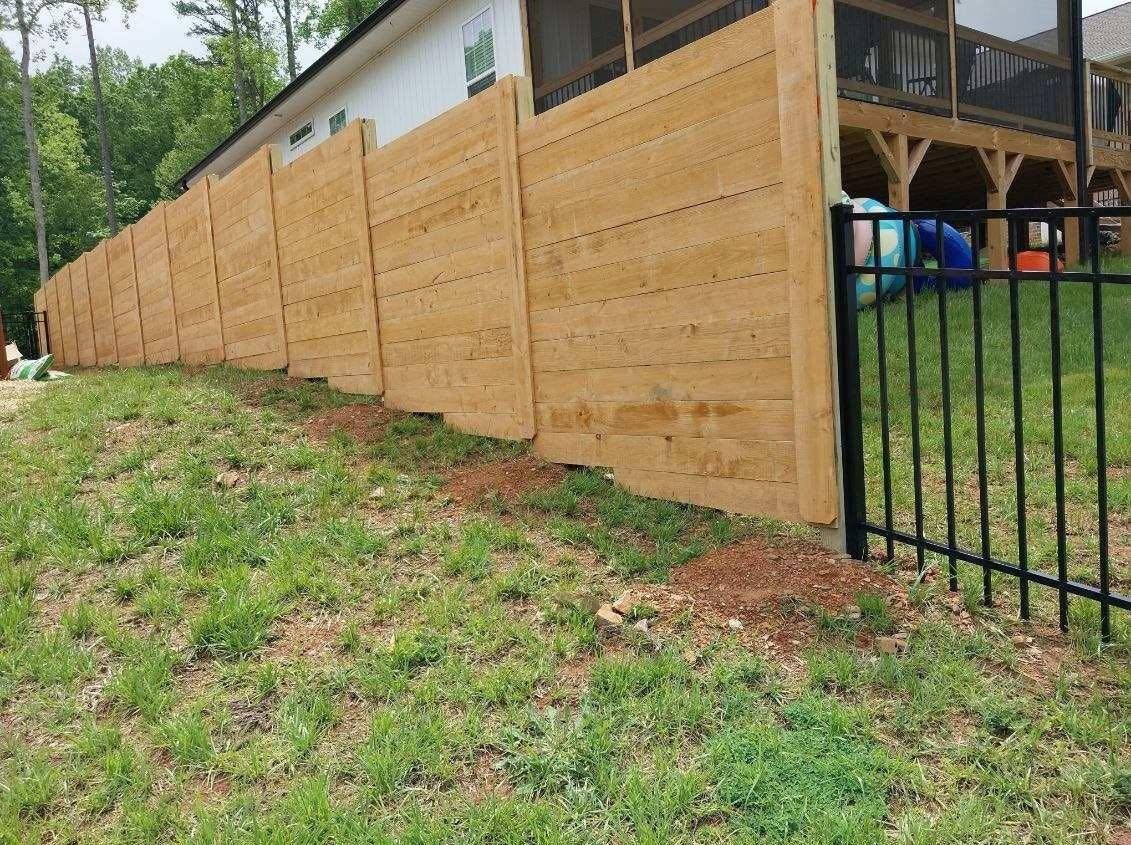 Wooden fence along a grassy slope, adjacent to a black metal fence and house.