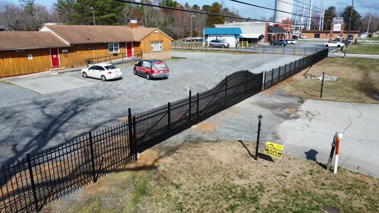 Black metal fence borders a gravel parking lot with cars parked near a low-slung building.