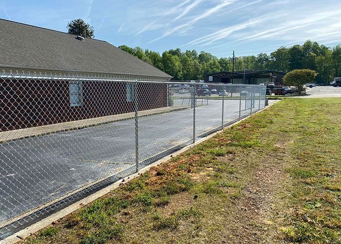 Chain-link fence borders a building with a dark roof and a parking lot on a sunny day.