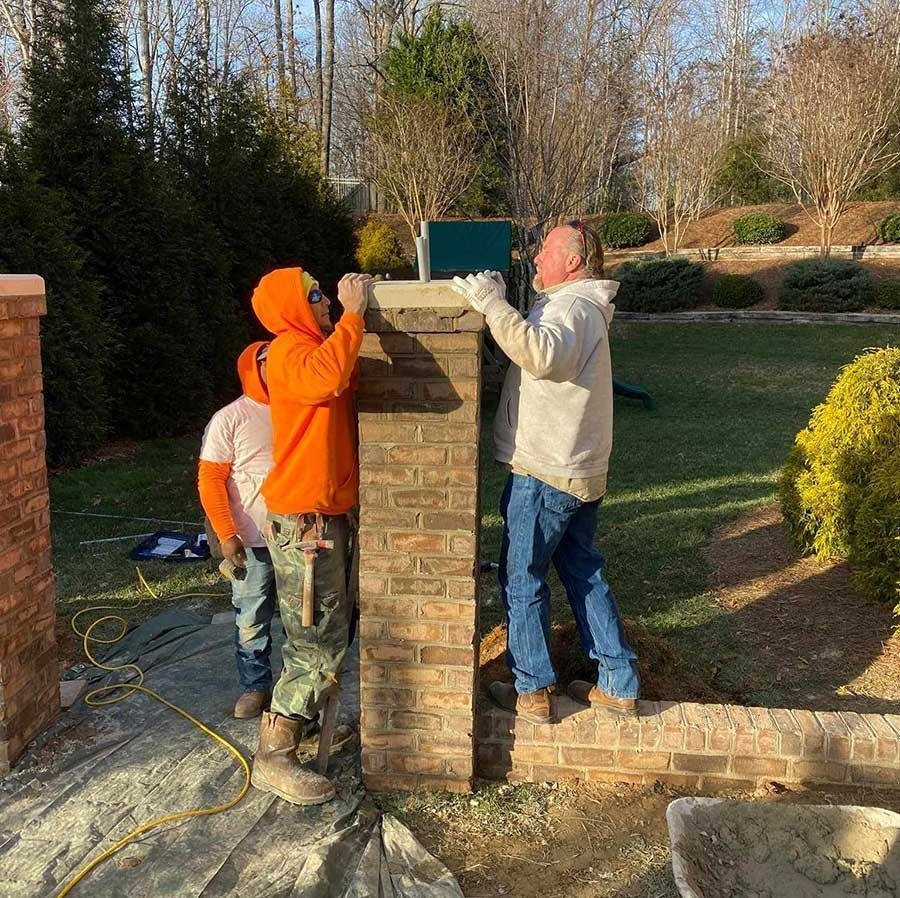 Three people constructing a brick pillar outdoors. Two are placing a capstone.