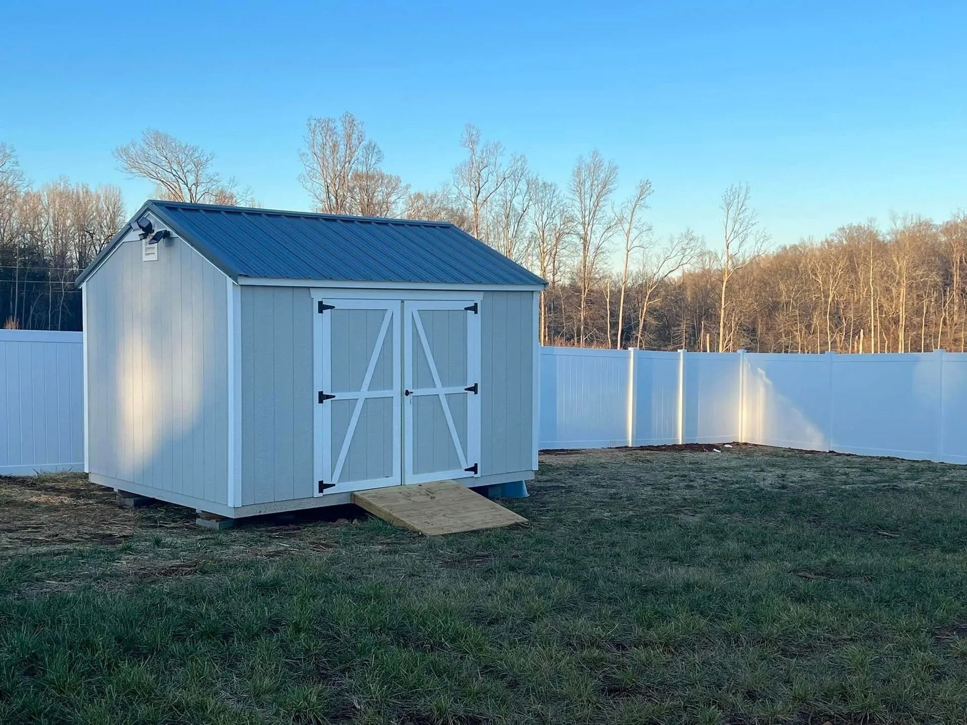 Light blue shed with white trim, blue roof, ramp, in a yard with a white fence.