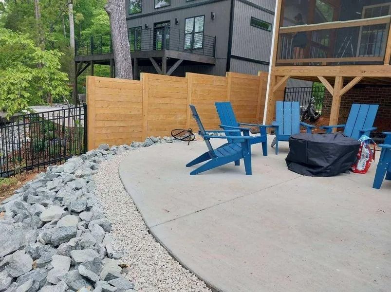 Patio with blue chairs around a fire pit, next to a wooden fence and rocky landscaping.