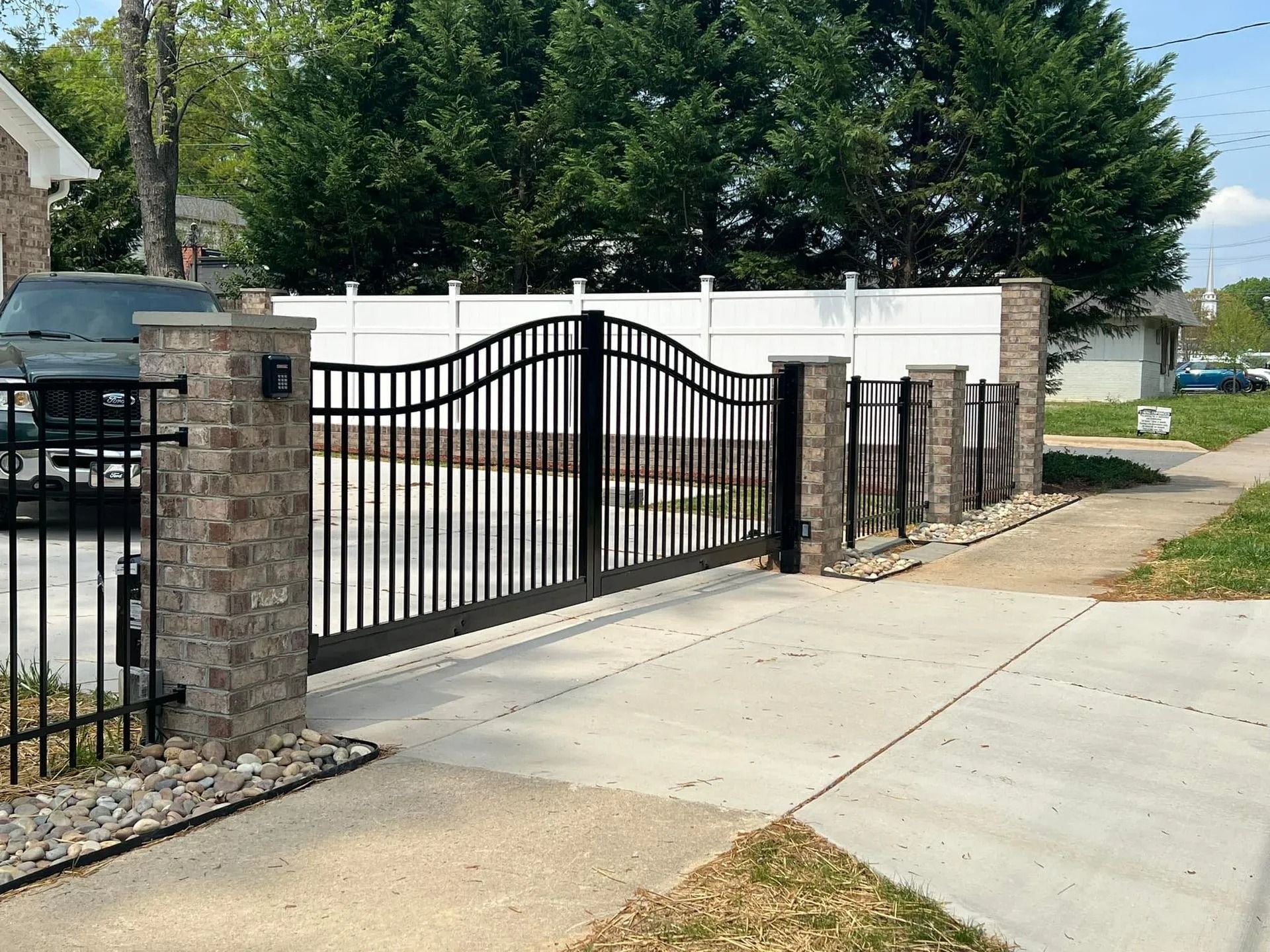 Black metal gate with stone pillars, leading to a driveway. White fence in the background, trees, and a car.