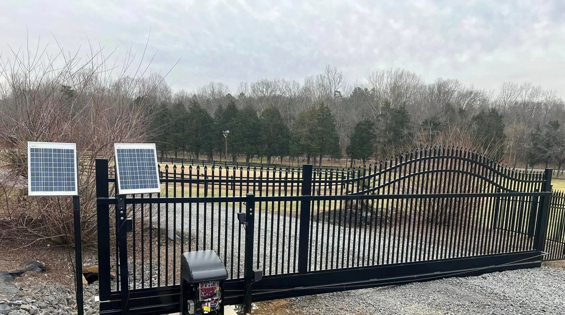 Black metal security gate with solar panels, gravel road, and trees in background. Cloudy day.