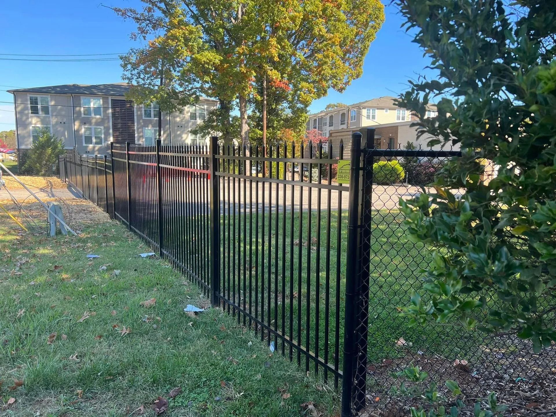 Black metal fence in front of a building and grassy area, with trees and a blue sky in the background.