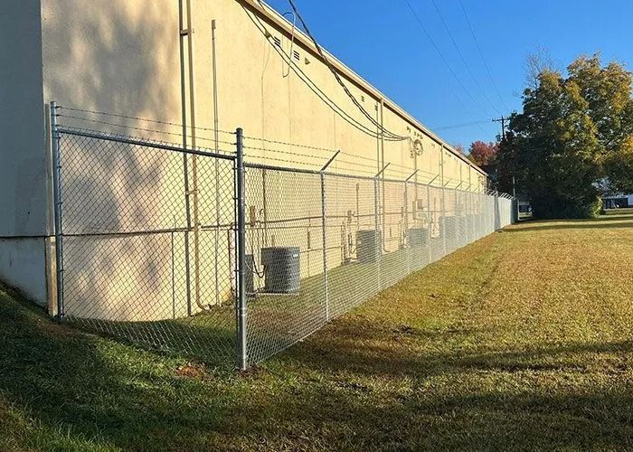 Chain-link fence with barbed wire surrounds a building with HVAC units. Sunny day, grassy area.
