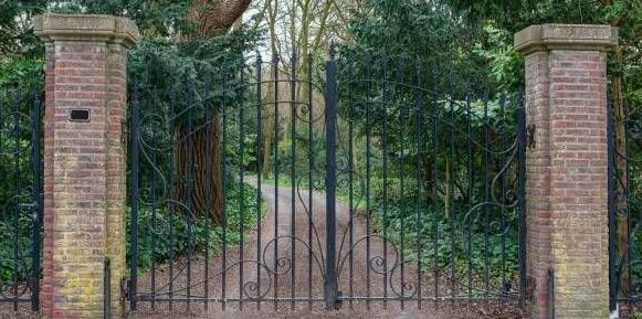 Wrought iron gate between brick pillars, leading to a path in a wooded area.