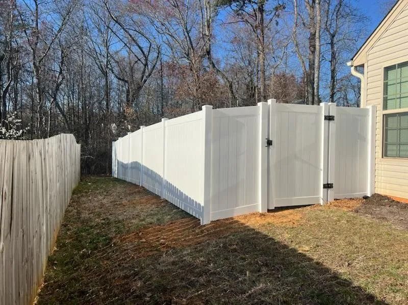 White vinyl fence curving through a grassy yard, partially obscuring a house and bare trees under a blue sky.