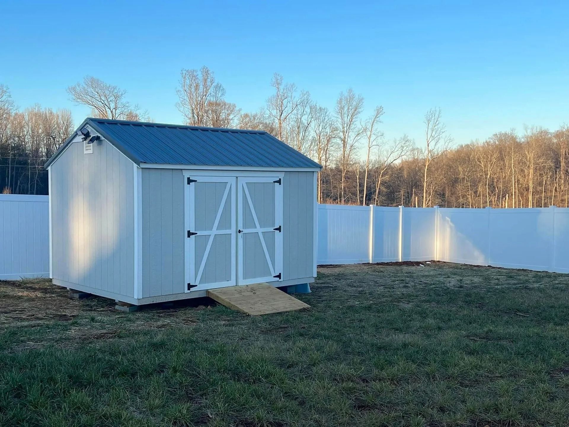 A light blue shed with a ramp and white fence, in a grassy backyard under a clear sky.