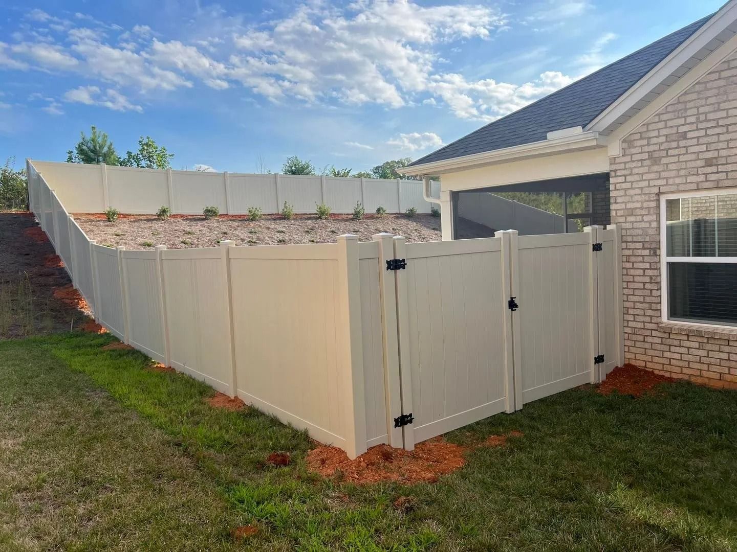 Beige vinyl fence surrounding a backyard with a gate, green grass, and a brick house.