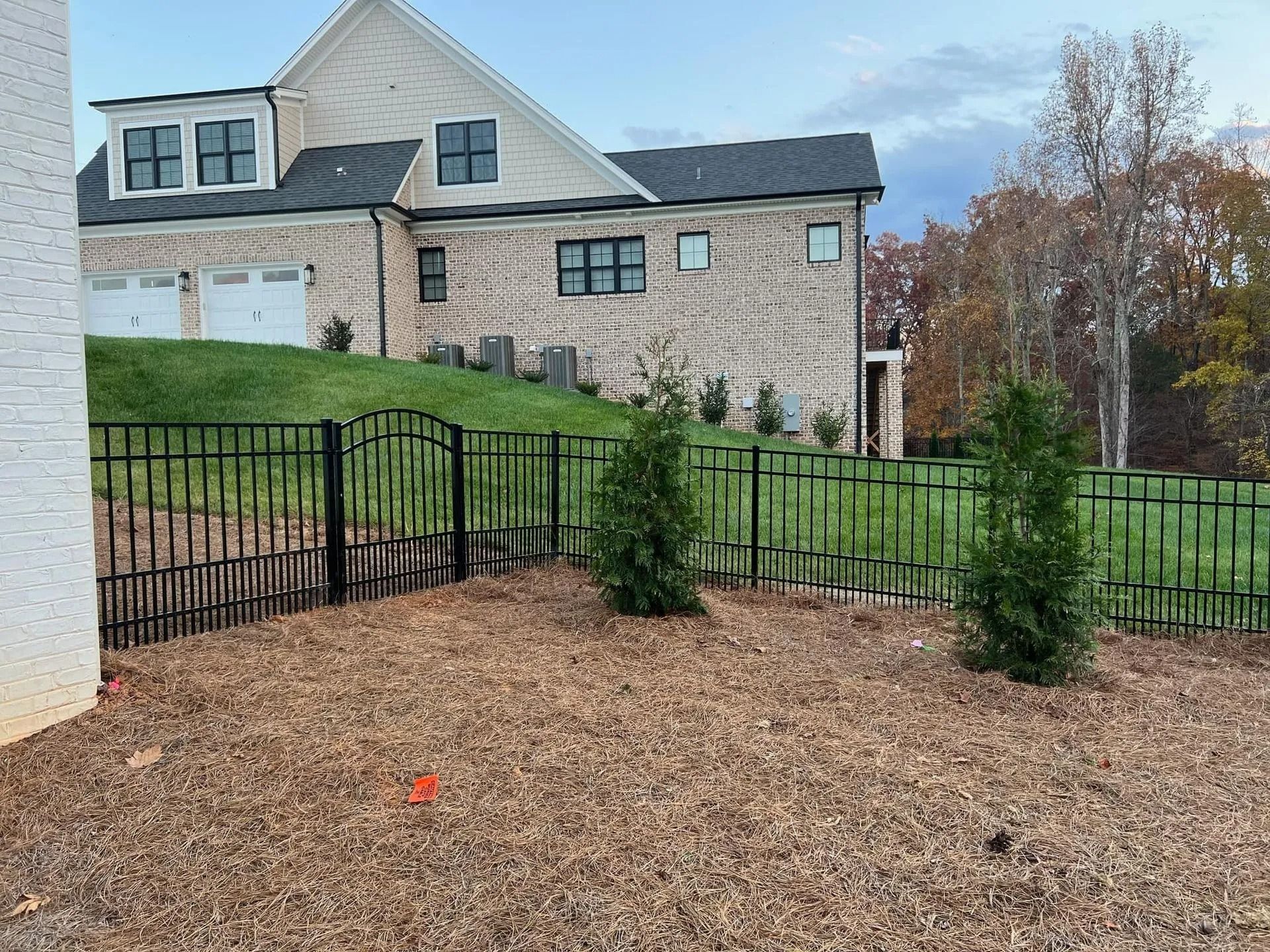 Black metal fence encloses a small yard with three green bushes. A large house is in the background.