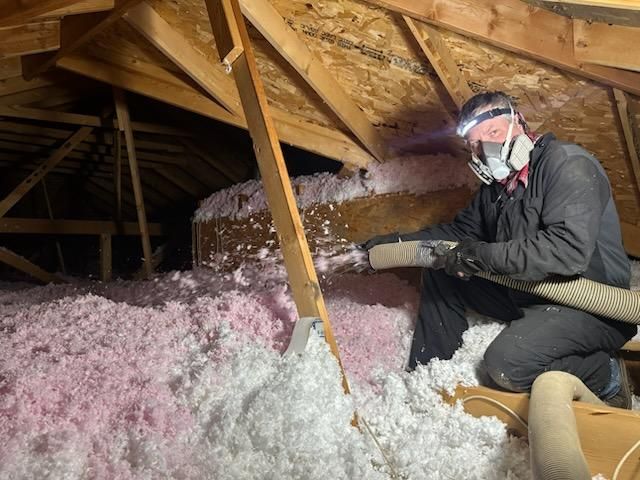 A person in an attic wearing a respirator and headlamp is blowing insulation.