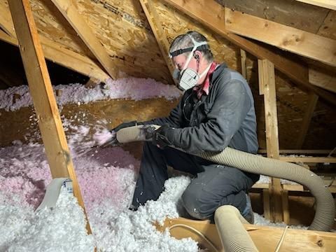 Person in an attic installing pink and white insulation, wearing a respirator.